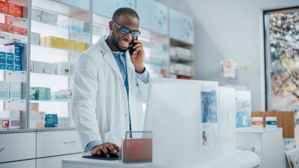 Pharmacy Drugstore Counter: Portrait of Helpful Black Male Pharmacist Talking on the Mobile Phone with Customer, Giving Medicine Recommendation, Uses Computer, Checks Prescription Drugs Availability Pharmacy Drugstore Counter: Portrait of Helpful Black Male Pharmacist Talking on the Mobile Phone with Customer, Giving Medicine Recommendation, Uses Computer, Checks Prescription Drugs Availability cashier photos stock pictures, royalty-free photos & images