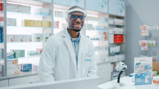 istock Pharmacy: Professional Confident Black Pharmacist Wearing Lab Coat and Face Shield, Looks at Camera Smiling Charmingly. Druggist in Drugstore Store with Shelves Medicine. Coronavirus Safety Measures 1325914378