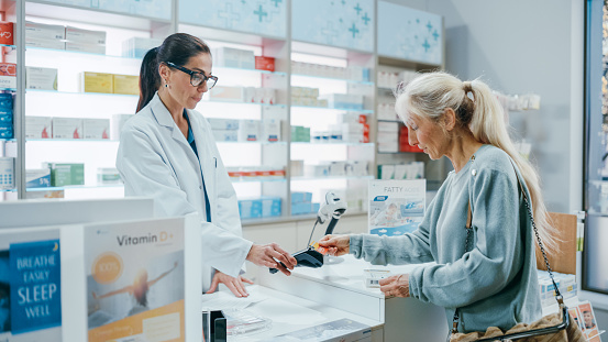 Pharmacy Drugstore Checkout Cashier Counter: Female Pharmacist Service Beautiful Senior Female Customer Buying Prescription Medicine Package and Paying Using Contactless Credit Card to Terminal