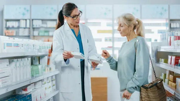 Photo of Pharmacy: Professional Specialist Pharmacist Helping Beautiful Senior Female Customer with Medicine Recommendation, Advice, Talking. Drugstore with Full of Drugs, Pills, Health Care, Beauty Products