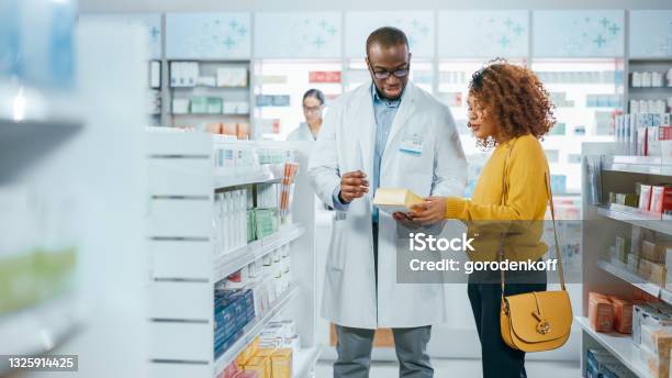 Pharmacy Professional Black Pharmacist Helping Beautiful Latin Female Customer With Medicine Recommendation Advice Talking Drugstore With Full Of Drugs Pills Health Care Beauty Product Packages Stock Photo - Download Image Now