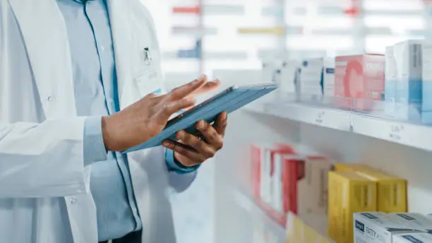 Photo of Pharmacy: Portrait of Professional Black Pharmacist Uses Digital Tablet Computer, Checks Inventory of Medicine, Drugs, Vitamins, Health Care Products. Druggist in Drugstore Store. Focus on Hands