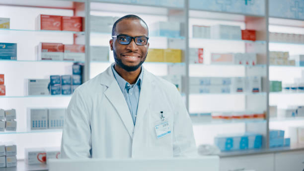 comptoir de pharmacie: portrait de beau pharmacien professionnel noir travaillant sur ordinateur, regarde la caméra souriant avec charme. spécialiste en pharmacie, étagères produits de soins de santé, médicament derrière - charmingly photos et images de collection