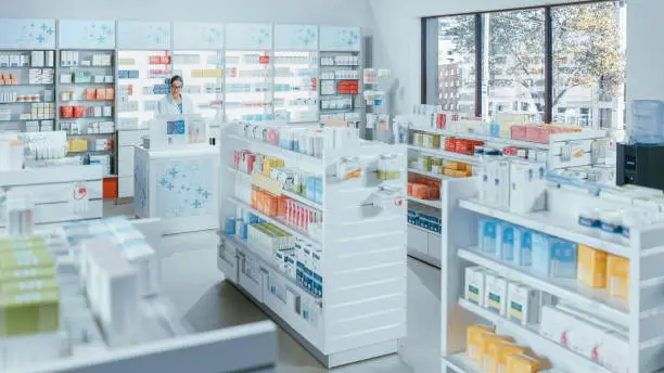 Photo of Modern Pharmacy Drugstore with Shelves full of Packages Full of Modern Medicine, Drugs, Vitamin Boxes, Supplements. In Background Professional Pharmacist Working at Checkout Counter.