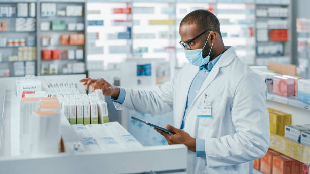 Pharmacy: Portrait of Professional Black Pharmacist Wearing Face Mask Uses Digital Tablet Computer, Checks Inventory of Medicine, Drugs, Vitamins, Health Care Products on a Shelf. Drugstore Store Pharmacy: Portrait of Professional Black Pharmacist Wearing Face Mask Uses Digital Tablet Computer, Checks Inventory of Medicine, Drugs, Vitamins, Health Care Products on a Shelf. Drugstore Store medical supplies stock pictures, royalty-free photos & images