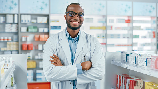Farmacia: Farmacéutico negro seguro profesional que usa abrigo de laboratorio y gafas, cruza brazos y mira a la cámara sonriendo encantadoramente. Droguería en la tienda de la farmacia con los productos del cuidado médico de los estantes photo