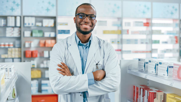 약국: 전문 자신감 검은 약사가 실험실 코트와 안경을 입고, 팔을 교차하고 매력적으로 미소 카메라를 찾습니다. 약국 매장에서 선반 건강 관리 제품 - pharmacy 뉴스 사진 이미지