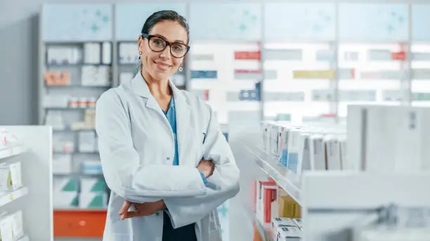 Photo of Pharmacy: Portrait of Beautiful Professional Caucasian Female Pharmacist Wearing Glasses, Crosses Arms and Looks at Camera Smiling Charmingly. Drugstore Store with Shelves Health Care Products