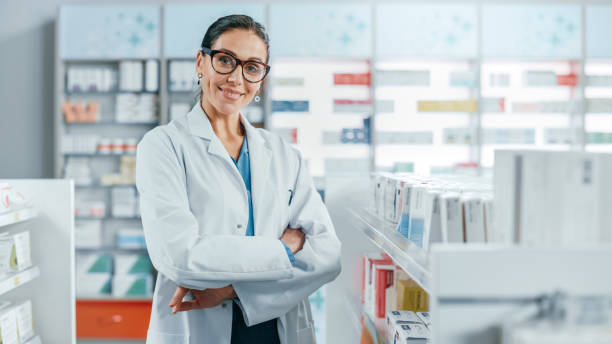 farmácia: retrato de linda profissional farmacêutica caucasiana usando óculos, cruzes braços e olhares para a câmera sorrindo encantadoramente. farmácia loja com produtos de saúde das prateleiras - lab coat women one person female - fotografias e filmes do acervo