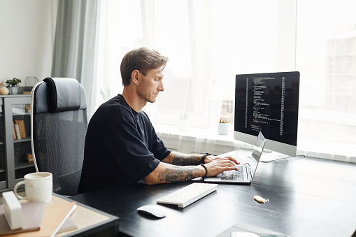 Young male programmer writing program code sitting at the workplace with monitor in the office