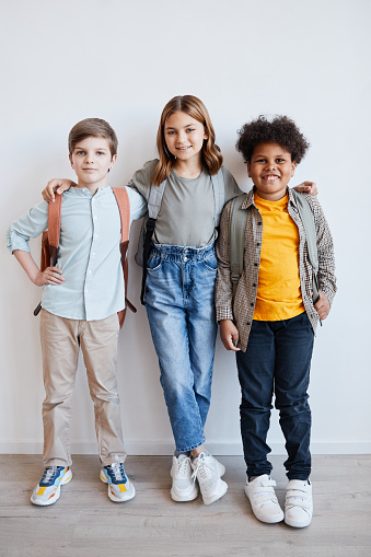 Full length portrait of three smiling schoolkids wearing backpacks and looking at camera while standing against white wall