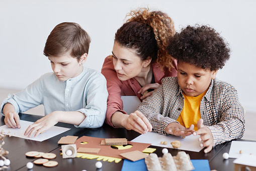 Portrait of two kids making cardboard models during art and craft class in school with female teacher
