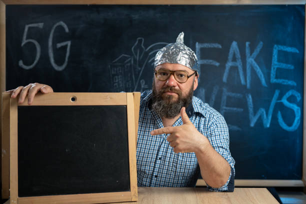 a strange guy conspiracy theorist in a protective aluminum foil hat and glasses sits at a table and points his finger at a whiteboard or space for text. fake news concept. - tin foil hat imagens e fotografias de stock