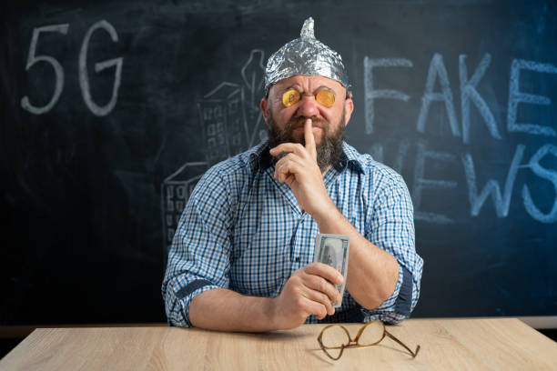 stupid conspiracy theorist with bitcoin coins in his eyes and dollar bills in his hand closes his mouth with his finger. secrets and fake news. conspiracy theory and currency. cryptocurrency. - tin foil hat imagens e fotografias de stock