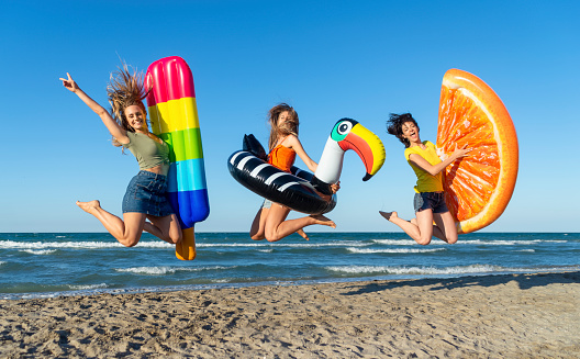 three happy girls on jumping motion with inflatable mattresses at the beach outdoors in summer celebrating vacation days. friends having fun on seaside holidays joy, carefree, genz and party concept