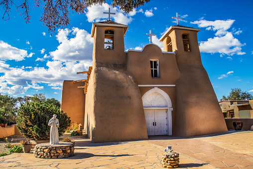 Historic adobe San Francisco de Asis Mission Church in Taos New Mexico in dramatic late afternoon light under intense blue sky with fluffy while clouds and birds in the belfry