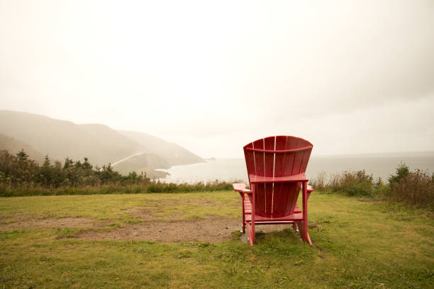 A single lounge chair sits on a grassy hill, overlooking a scenic view of green fields and trees under a clear blue sky. The setting exudes tranquility, inviting relaxation amidst nature.