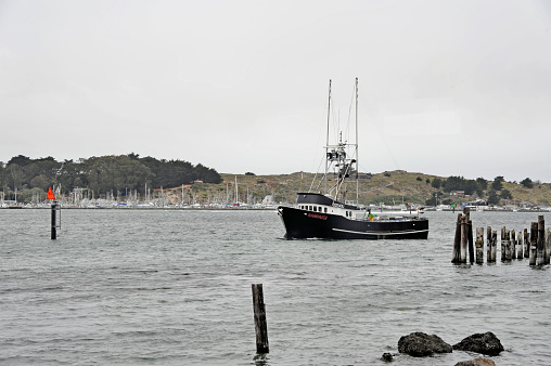 A fishing boat with a fisherman at the stern who is returning to the port with mountains as a backdrop - Black and white