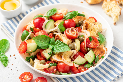 Panzanella - Italian salad from vegetables and bread. Salad with tomatoes, onions, cucumbers, bread, capers and basil in a white plate on a gray concrete background. Vegan salad, Italian cuisine