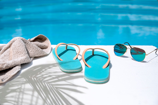 Towel, flip flops and sunglasses on the white poolside on turquoise pool water. Tropical pool time background Copy space.