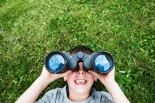 A young Caucasian boy lays on a green grass covered yard and looks up at the sky with an old worn pair of binoculars.  A big smile is in on his face.