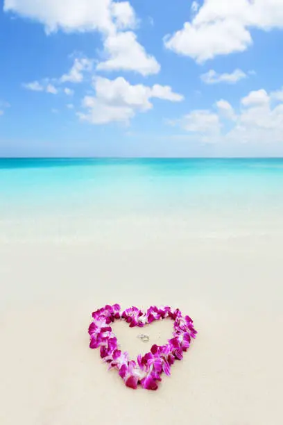 Photo of Two wedding rings in a heart lei on beach vacation. Hawaiian flower necklace lying on the sand in the shape of a heart with rings for marriage proposal in tropical vacation destination honeymoon.