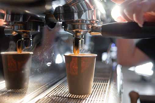 Close-up of a coffee machine making an espresso. The coffee flows into a paper cup under the metal spout of the coffee maker. Production of hot coffee in disposable containers.