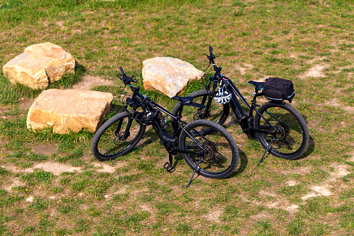 Two electric bicycles stand in a meadow near the stones. Top view. Bicycle helmets hang on the handlebars.