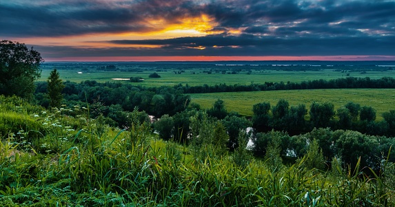 Beautiful dramatic sunset over a river valley with green meadows. Panoramic image. View from the hill