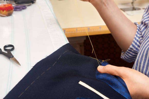istock Woman tailor wrapped up in sewing jacket with needle and thread while sitting at working table. Suit tailoring in process of custom-made jacket. Bespoke suit tailoring. 1325957927