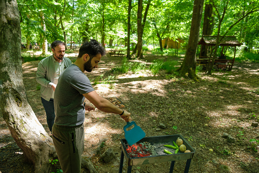 a guy stokes fire up with vegetables cooking in public picnic forest place and another friend comes for help
