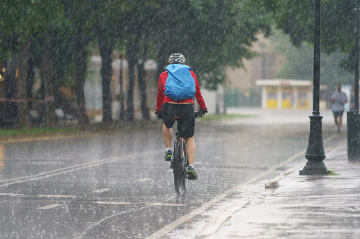 Moscow, Russia. June 27, 2021: Raining day in city at summer. Wet young man ceycling. Texture of strong, fresh and powerful water drops and sprays. Tropical storm as result of global climate warming. Back, rear view.