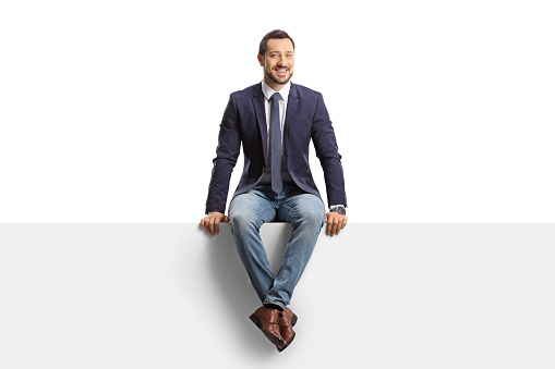 Full length portrait of a man in jeans and suit sitting on a blank panel isolated on white background
