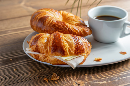 Two French croissants with ham and cheese and a cup of black coffee on the white plate. Wooden table, morning country breakfast. Side view at an angle.