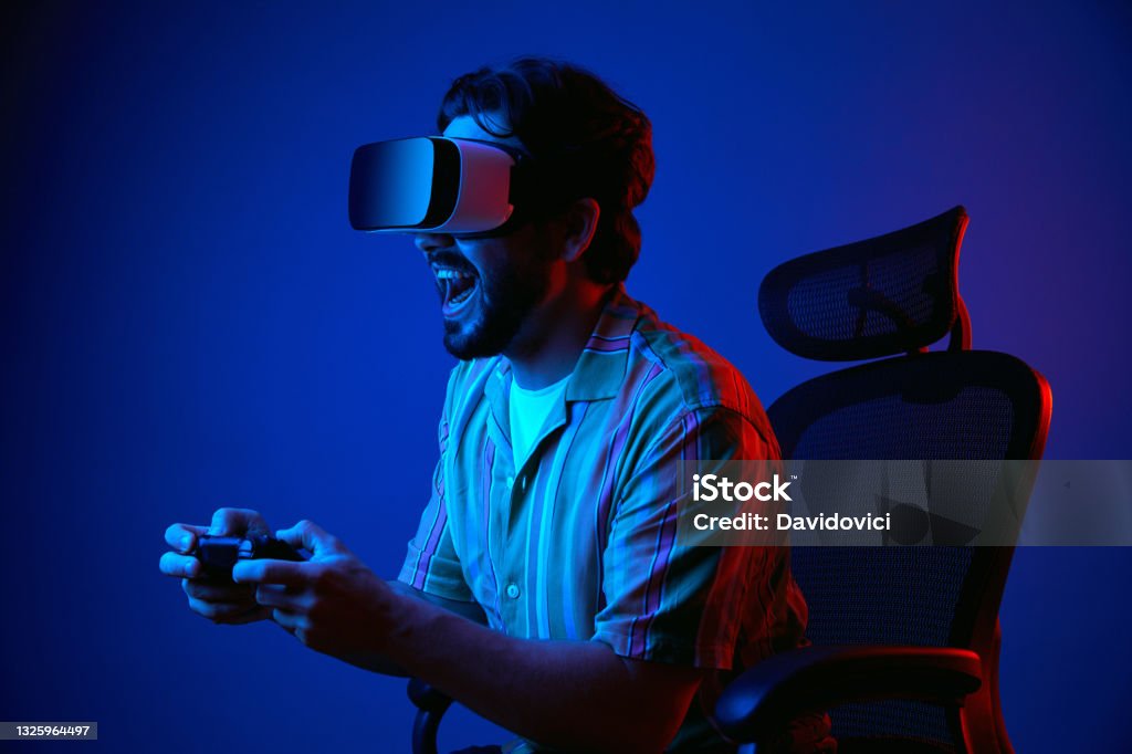 Man screams and plays with joystick in VR glasses while sitting on chair Young bearded man screaming and playing game with joystick in VR glasses while sitting on chair against dark blue background Gamer Stock Photo