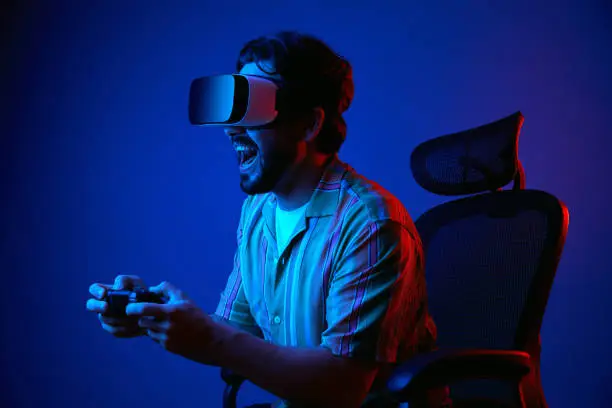 Young bearded man screaming and playing game with joystick in VR glasses while sitting on chair against dark blue background