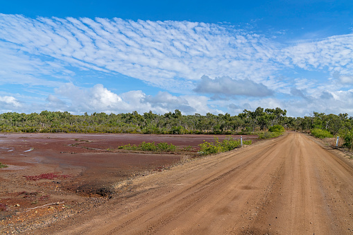 Dirt road beside salt flats and bushland under a cloudy blue sky