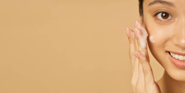 istock Website header of Studio half face portrait of beautiful young woman smiling at camera while applying gentle foam facial cleanser isolated over beige background 1325979559