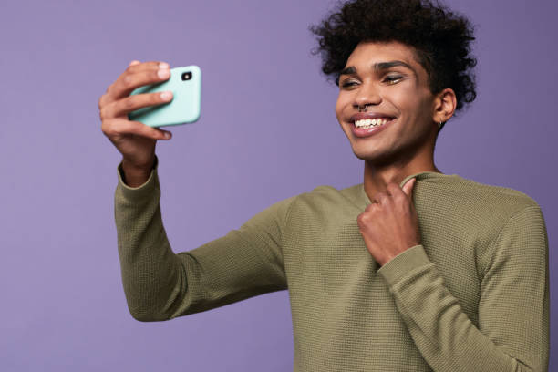 Portrait of transgender latino male taking selfies with mobile in hand. Happy trans gender young man Portrait of transgender latino male taking selfies with mobile in hand. Happy trans gender young man calling via video on phone and smiling. Fashion studio shot on purple background transgender-athletes stock pictures, royalty-free photos & images
