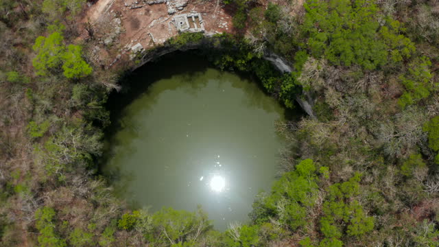 Forwards tilt-down reveal of large round Cenote sinkhole in ground. Sun reflecting on water surface. Historical monuments of pre-Columbian era, Chichen Itza, Mexico