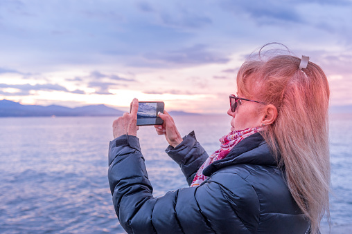 Adult blonde woman using her smart-phone to take a photo of the coast during sunset.
