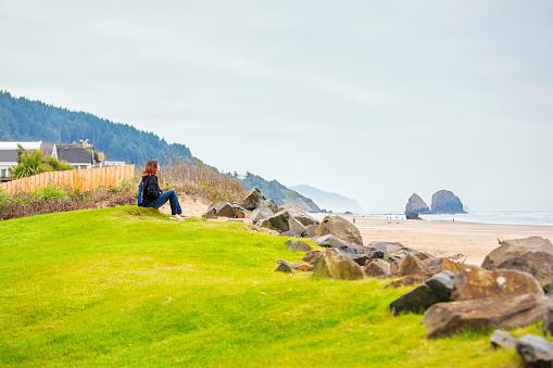 One teen girl with dyed red hair sitting alone on grassy hill next to beach looking out towards ocean on foggy day
