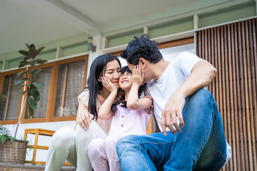 Asain happy family spend leisure time together, Parents kiss on young little girl daughter's cheek with gentle in garden in front of house. Loving Father and moter enjoy parenting and activity at home