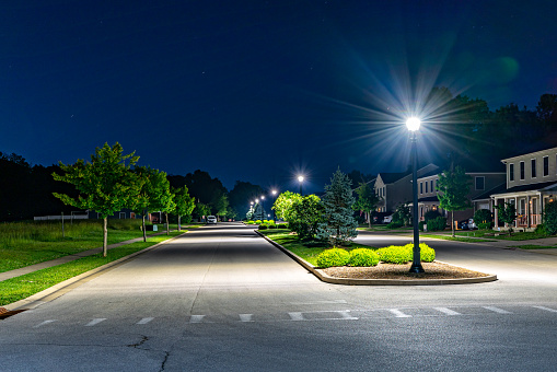 Neighborhood street at nighttime with street lights and plants on the side of the road and a traffic island in the middle.