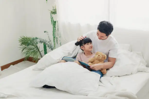 Photo of Happy Asian dad with cute daughter holding reading book together in the bed room, smiling father and kid girl having fun being at home spending time together, friendly parents and child readers concept