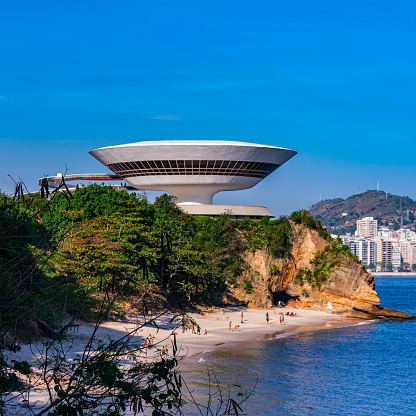 Rio de Janeiro, Brazil - June 8, 2023: Niemeyer Museum of Contemporary Arts. An ocean bordered by rocky cliffs and trees. A white bowl-shaped building