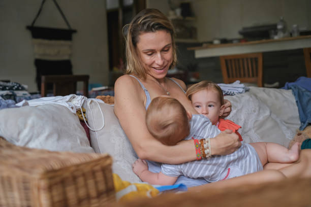 Shot of a caring Caucasian mother breastfeeding her son on the sofa accompanied by her first-born daughter who is leaning to her side.