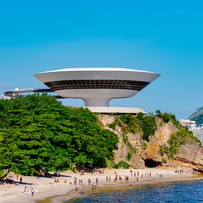 Rio de Janeiro, Brazil - June 8, 2023: Neimeyer Museum of Contemporary Art. A bowl-shaped structure beside the sea. People walk and gather around this architectural landmark.