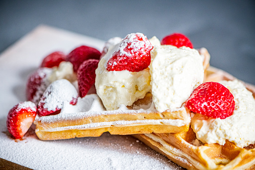 Waffels with whipped cream and strawberries topped with icing sugar. The homemade waffels are presented on a wooden board with a rustic look.