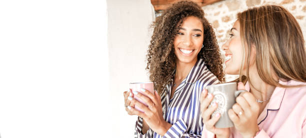 coppia di donne multirazziali sorridenti che si guardano tenendo una tazza da tè in pigiama si svegliano. nuove normali relazioni e abitudini di vita quotidiana delle famiglie gay. donna riccia bruna afro-americana - coffee couple italy drinking foto e immagini stock